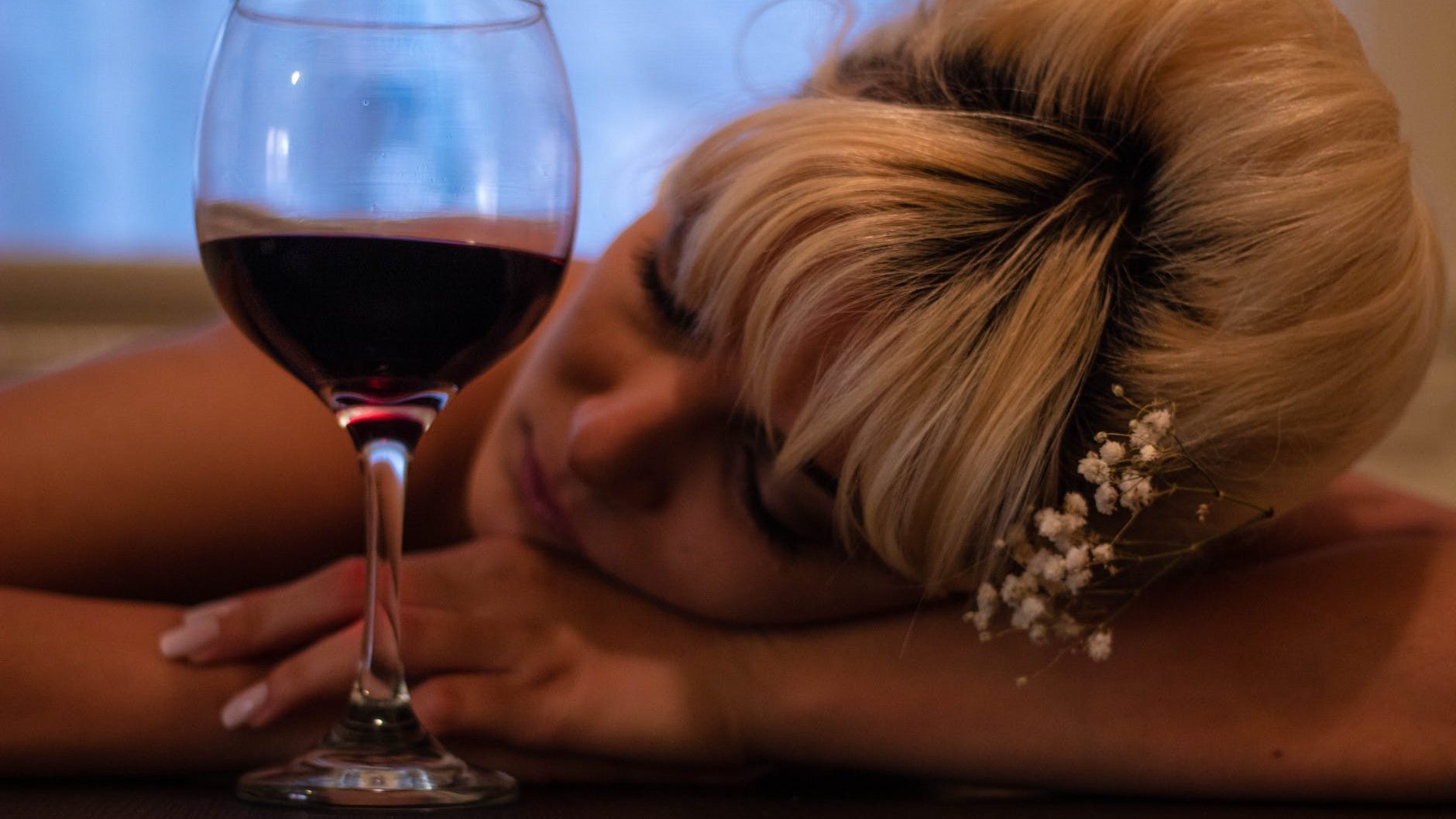 woman with white flower accent headdress leaning her head on table beside half filled wine glass